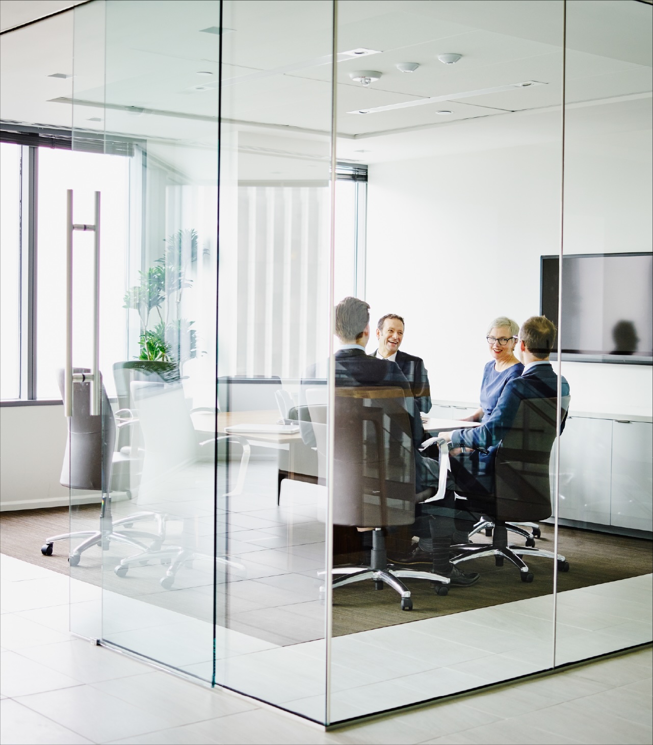 Business meeting in a modern glass-walled conference room, with four people seated around a table, collaborating in a bright, minimalist office setting.